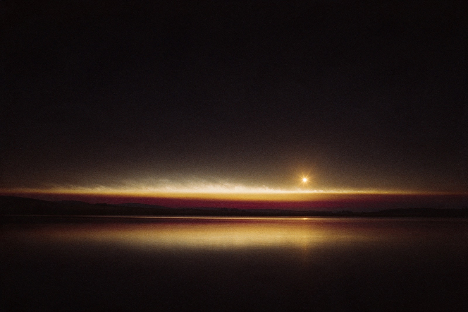 Ein heller Lichtpunkt knapp über dem Horizont, darunter ein goldroter Dämmerungsstreifen und ein stiller See, in dem sich das Licht spiegelt.